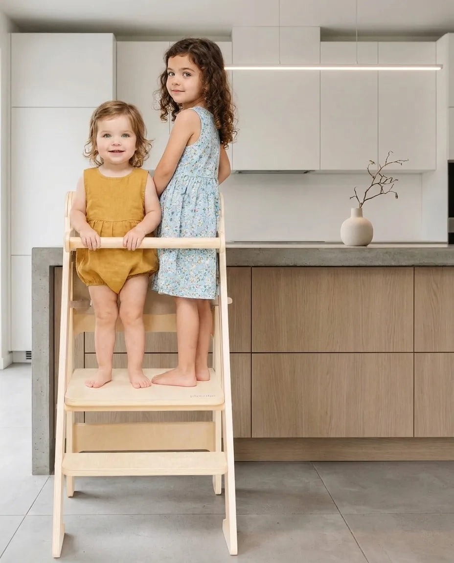 Two children standing on a wooden ikiddoo learning tower beside a kitchen counter