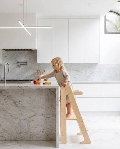 Child on a learning tower reaching for fruit in a bright kitchen