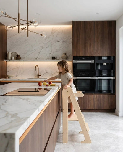Child on a learning tower reaching for fruit in a warm kitchen