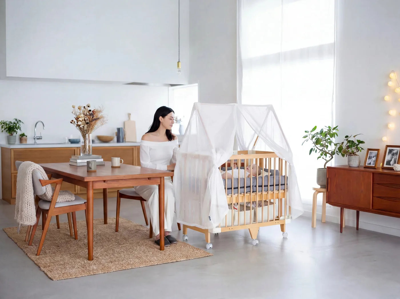 A mother drinks coffee at the table while her baby sleeps in the Creative Cot nearby